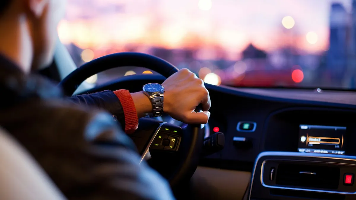 A driver's hand on the steering wheel at dusk, with city lights blurred ahead