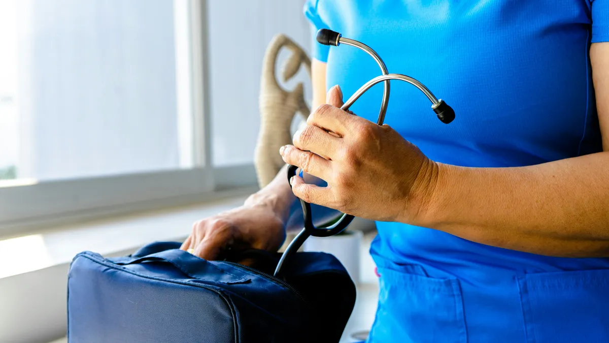 A nurse in blue scrubs packs a stethoscope into a navy travel bag beside a bright window