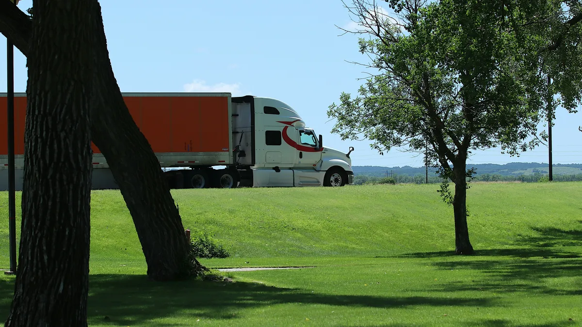 Camión semirremolque blanco con tráiler naranja por una carretera, junto a pasto verde y árboles